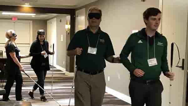 2 pictures show people traveling using human guide in a large hallway.  In the background are 2 men, with the one being guided under blindfold with a cane held parallel to his body, and the man guiding has his blindfold pushed to his forehead.  IN the foreground, although 2 people are pictured, the person providing human guide is mostly out of frame, with only their elbow and hip showing.  The person being guided has a blindfold on and a long cane extended.